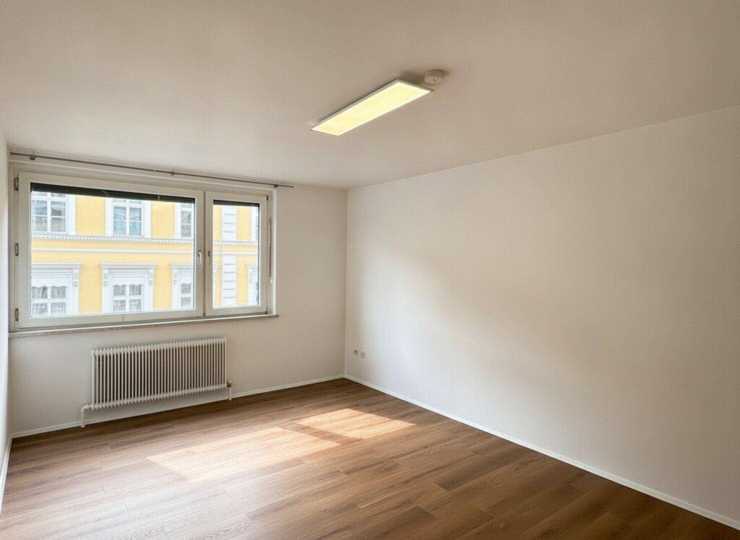 Heller, leerer Raum mit Holzboden, großem Fenster und Blick auf die gegenüberliegende Fassade, gut beleuchtet.