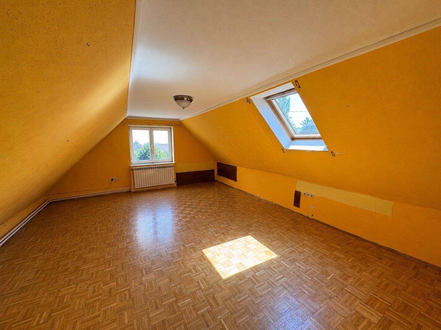 Helles Dachzimmer mit gelben Schrägen, Parkettboden und zwei Fenstern, darunter ein Dachfenster.