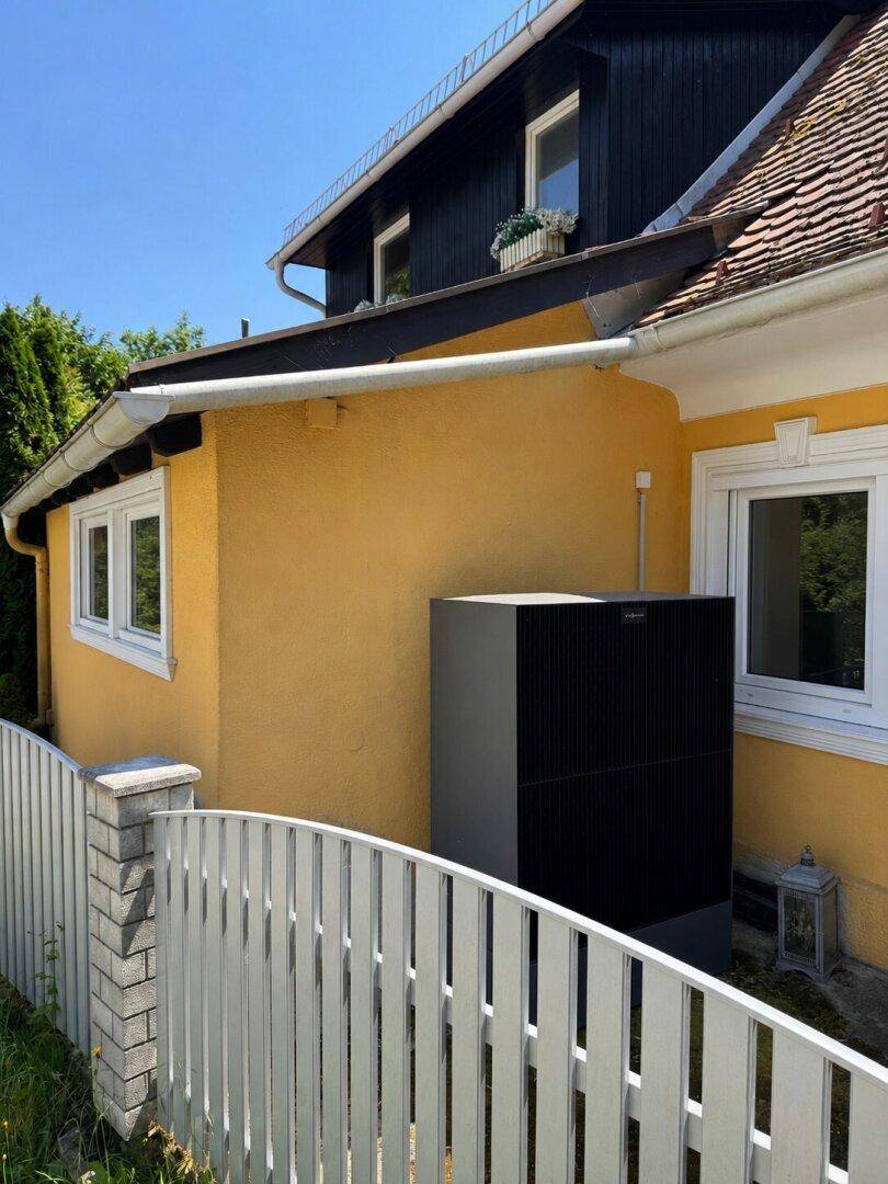 Moderne dunkle Wärmepumpe an der gelben Hauswand, hinter einem weißen Zaun, unter blauem Himmel.
