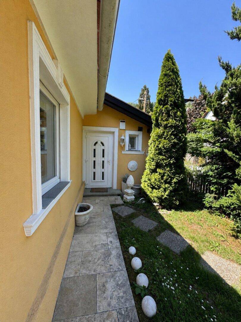 Eingangsbereich des Hauses mit weißer Tür, Steinweg und gepflegtem Rasen, umgeben von hohen Bäumen.