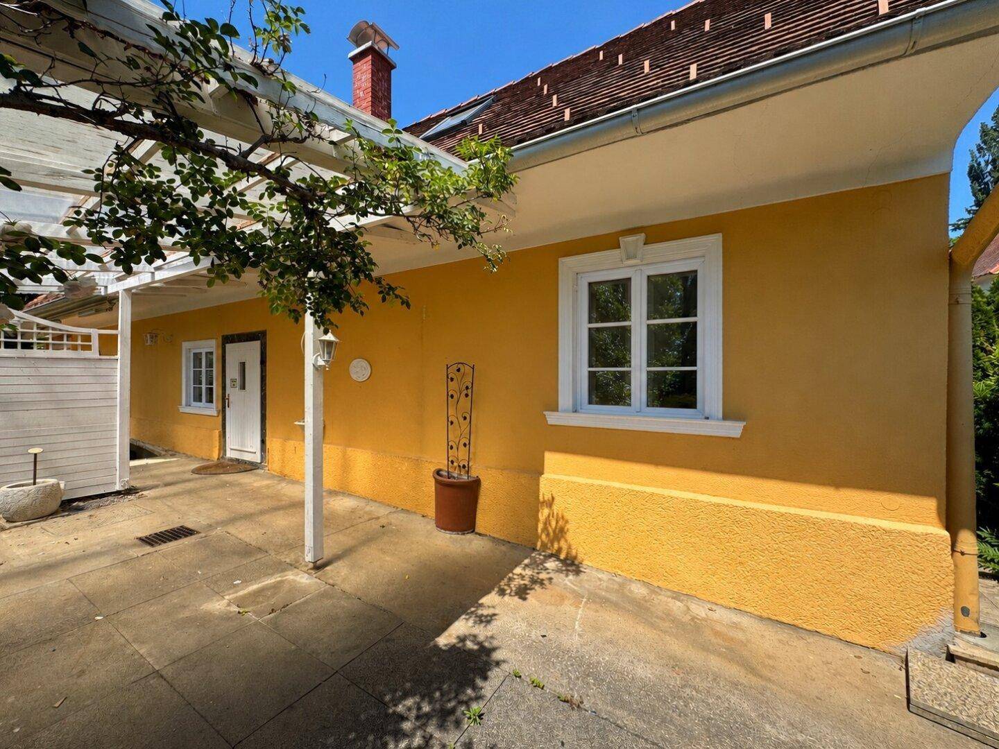 Überdachte Terrasse am gelben Haus mit Kletterpflanzen und gepflastertem Boden, ideal für Entspannung im Freien.