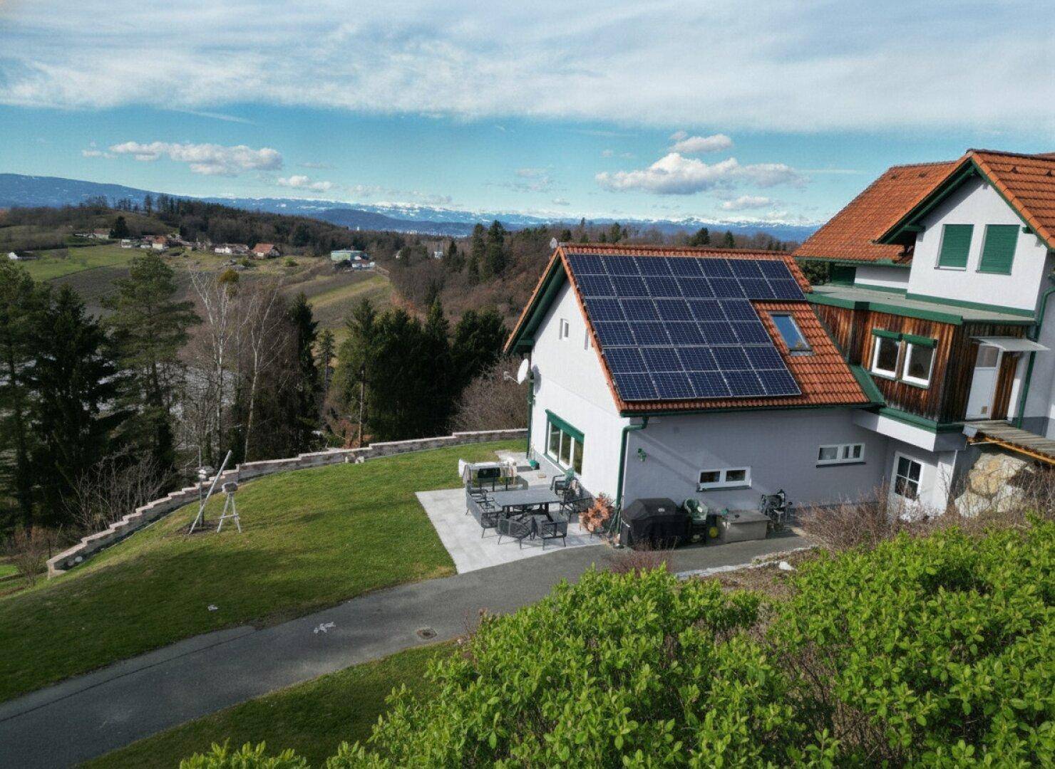Hausansicht mit Terrasse, Esstisch im Freien und Solaranlage auf dem Dach, umgeben von Grün.
