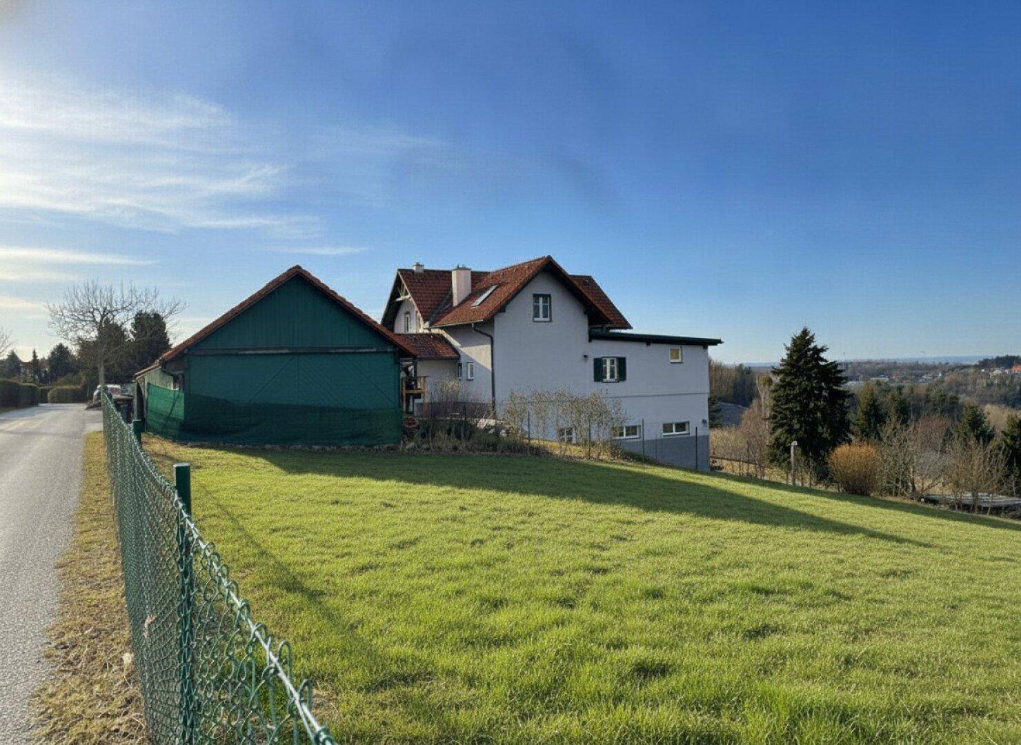Haus mit Anbau und weitläufiger Rasenfläche, umgeben von grüner Landschaft und Fernblick.