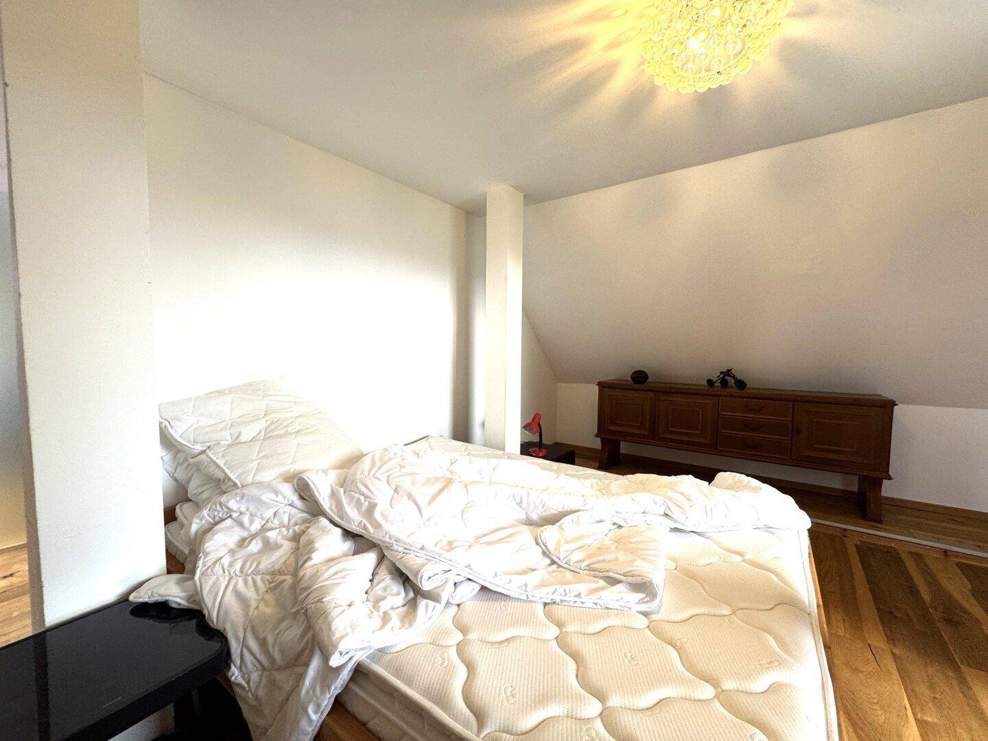 Schlafzimmer mit Bett, Kommode und schräger Decke, hell und einfach eingerichtet.