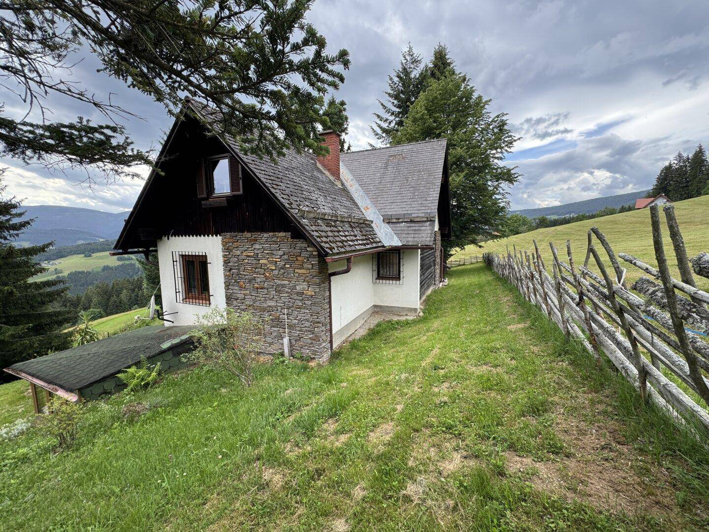 Kleines Nebengebäude mit Steinfassade und Holzzaun in ländlicher Umgebung, umgeben von Wiesen.