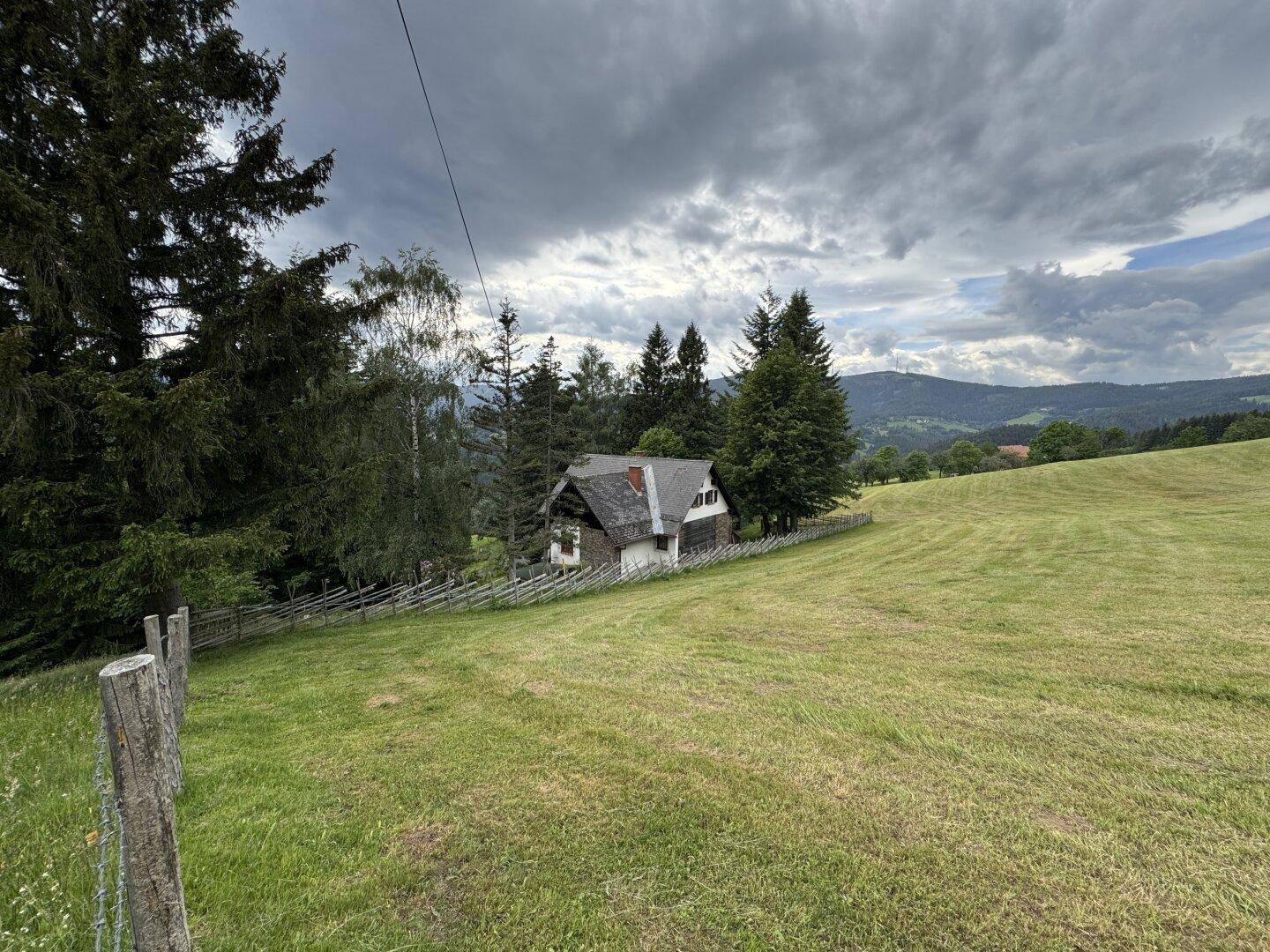Weitläufige Ansicht des Hauses in einer offenen Wiesenlandschaft mit bewaldeten Hügeln im Hintergrund.