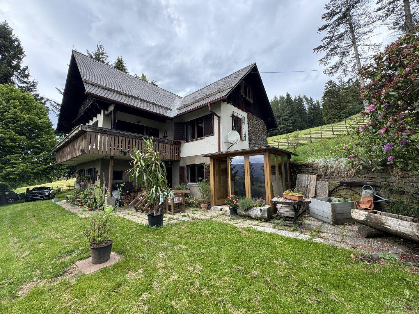 Traditionelles Haus mit Holzbalkonen und einem kleinen Wintergarten, umgeben von Grünflächen und Bäumen.