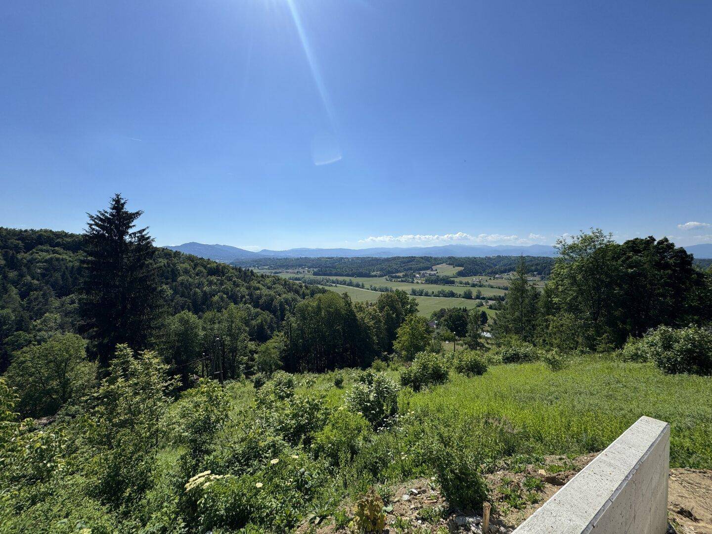 Panoramablick über die hügelige, bewaldete Landschaft mit weiten Feldern und Bergen am Horizont.