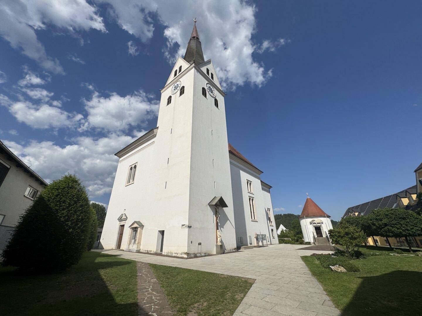 Hohe weiße Kirche mit spitzem Turm und Uhr, umgeben von Grünflächen und blauem Himmel.