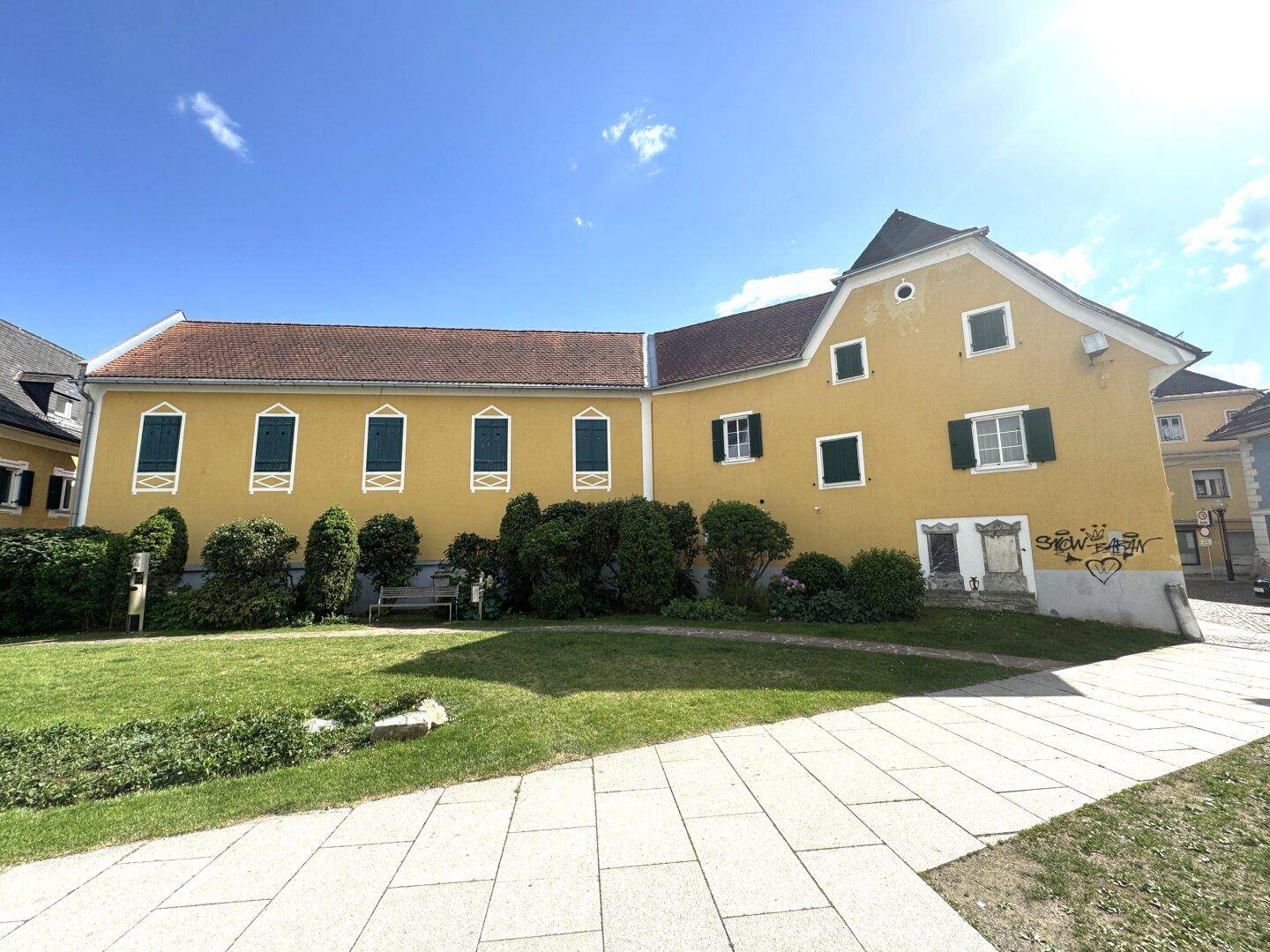 Langes gelbes Gebäude mit grünen Sträuchern und Rasenfläche unter sonnigem Himmel.