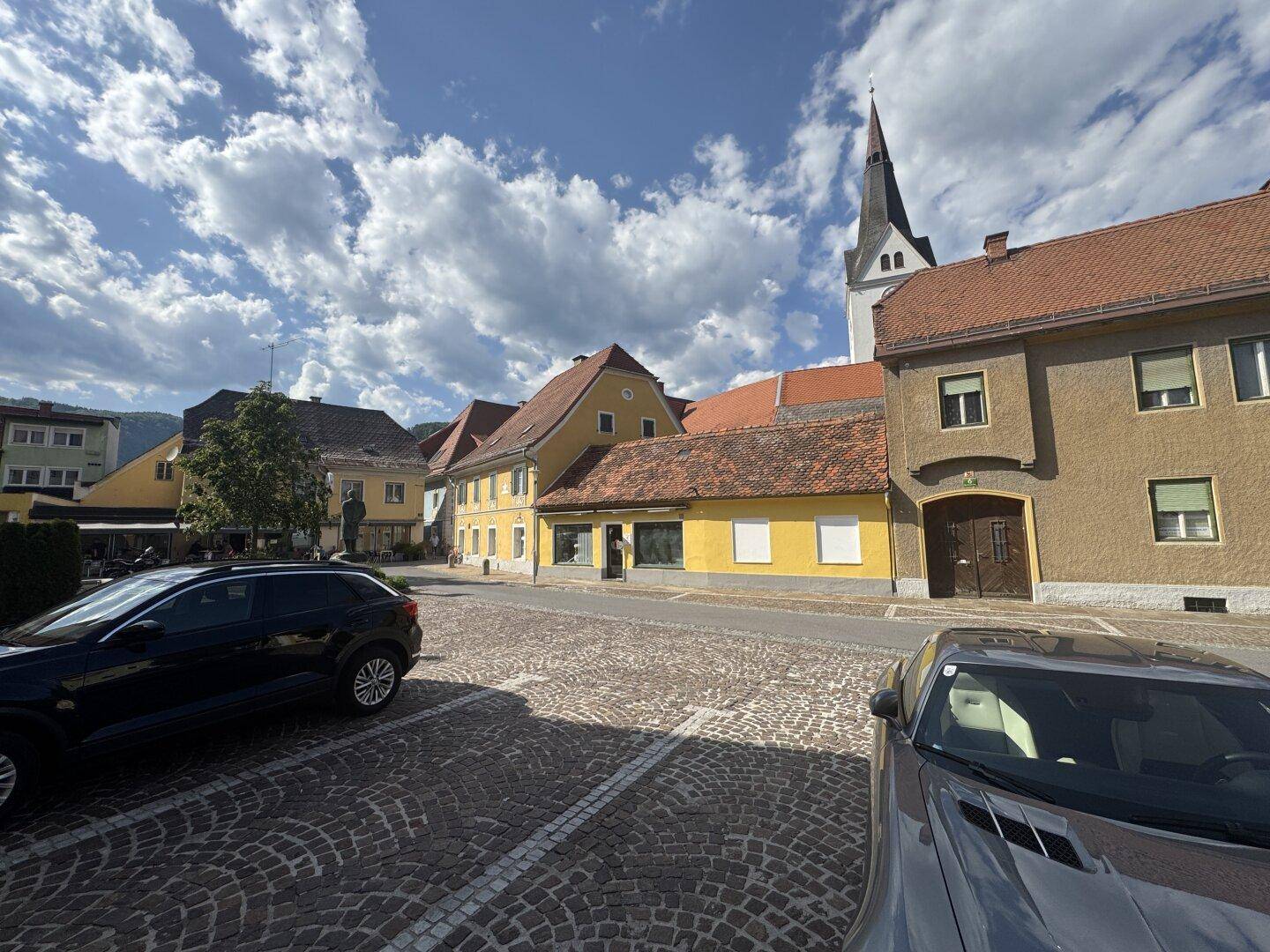 Kopfsteinpflasterstraße mit geparkten Autos und historischen Gebäuden, Kirche im Hintergrund.