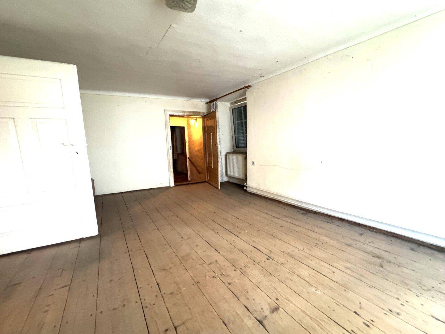 Leerer Raum mit Holzdielenboden, weißen Wänden und einem Fenster mit Heizkörper.