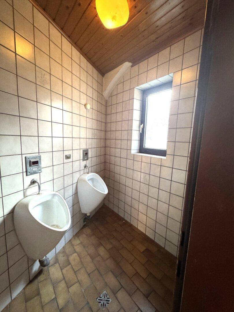 Gefliester Toilettenraum mit zwei Urinalen, Holzdecke und einem Fenster.