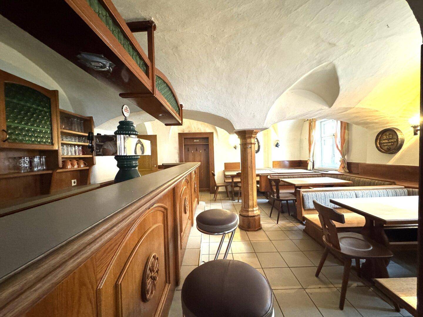 Rustikaler Gastraum mit Holzbalken, Bar und traditionellen Sitzbänken, gefliestem Boden.