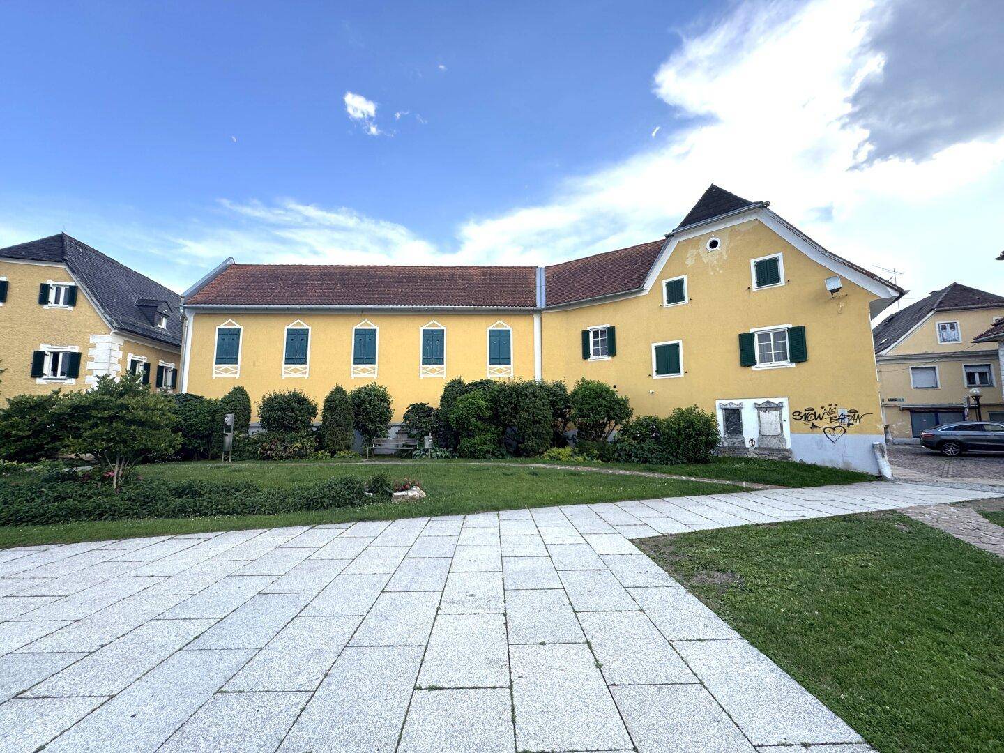 Traditionelles gelbes Gebäude mit grünen Fensterläden und gepflegtem Rasen unter blauem Himmel.