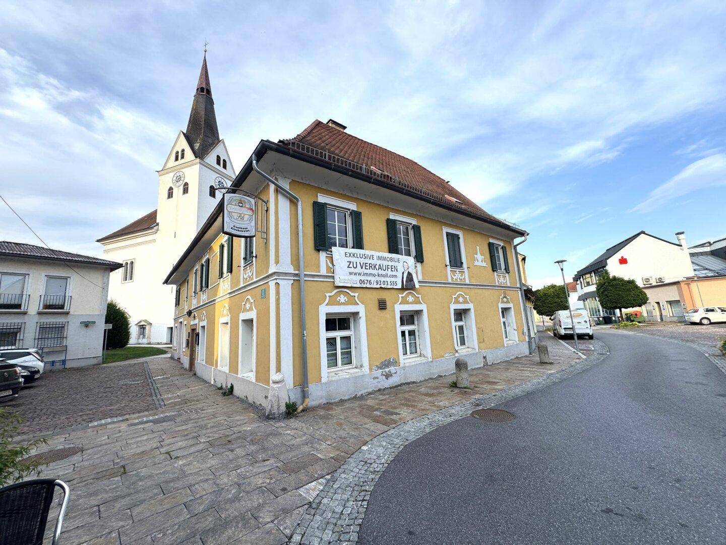 Historisches gelbes Gebäude an einer Straßenecke mit Kirche im Hintergrund und "Zu Verkaufen" Banner.