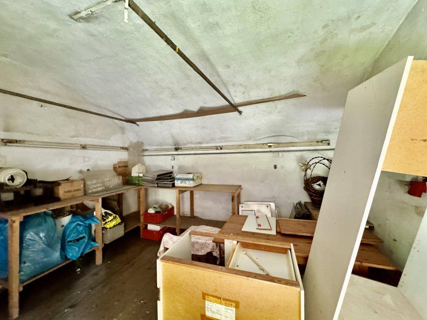 Unaufgeräumter Abstellraum mit Holztischen, verschiedenen Gegenständen und einer einfachen Decke.