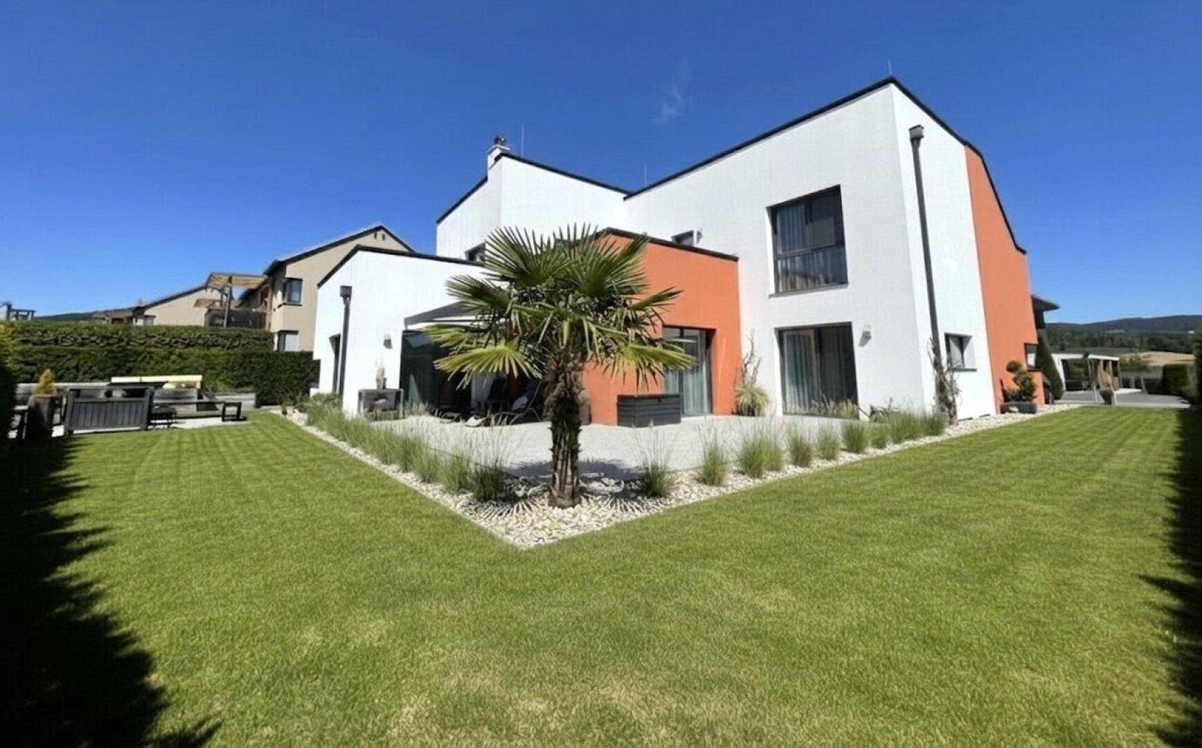 Moderne Hausfassade mit weißem und orangefarbenem Putz, umgeben von einem gepflegten Rasen und einer Terrasse.
