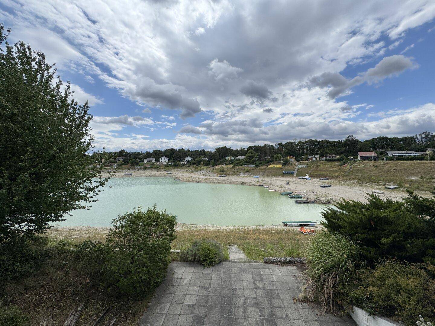 Panoramablick auf einen See mit Uferbereich und umliegender Natur, gesehen von einer gepflasterten Fläche.
