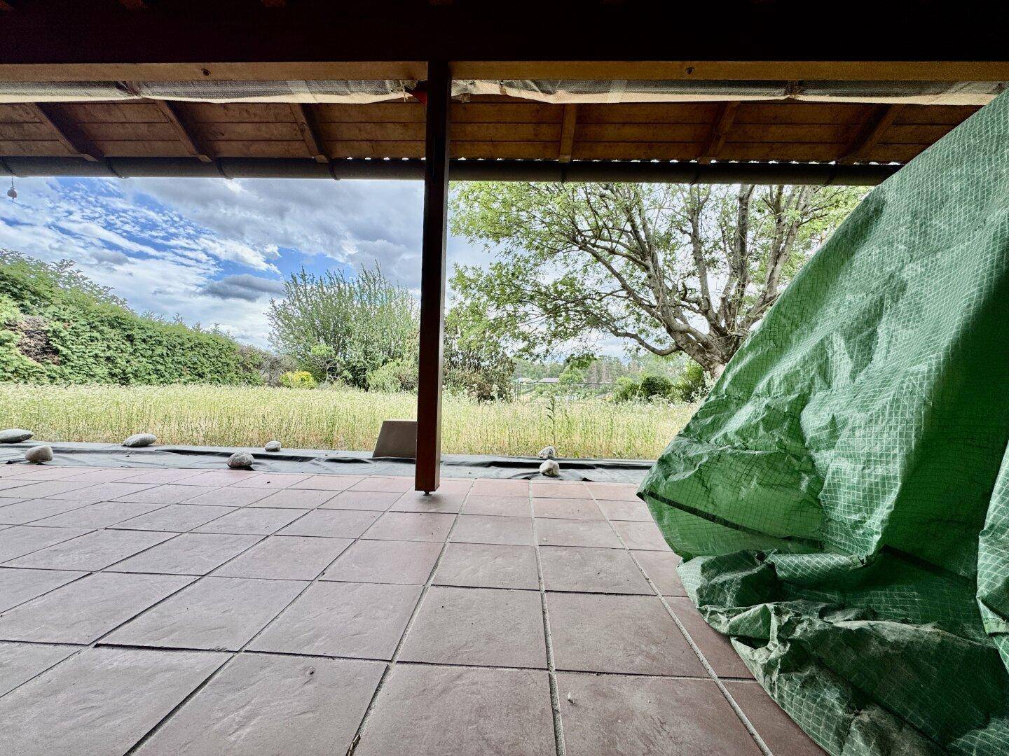 Überdachte Terrasse mit Fliesenboden und Blick auf eine grüne Wiese und Bäume im Hintergrund.