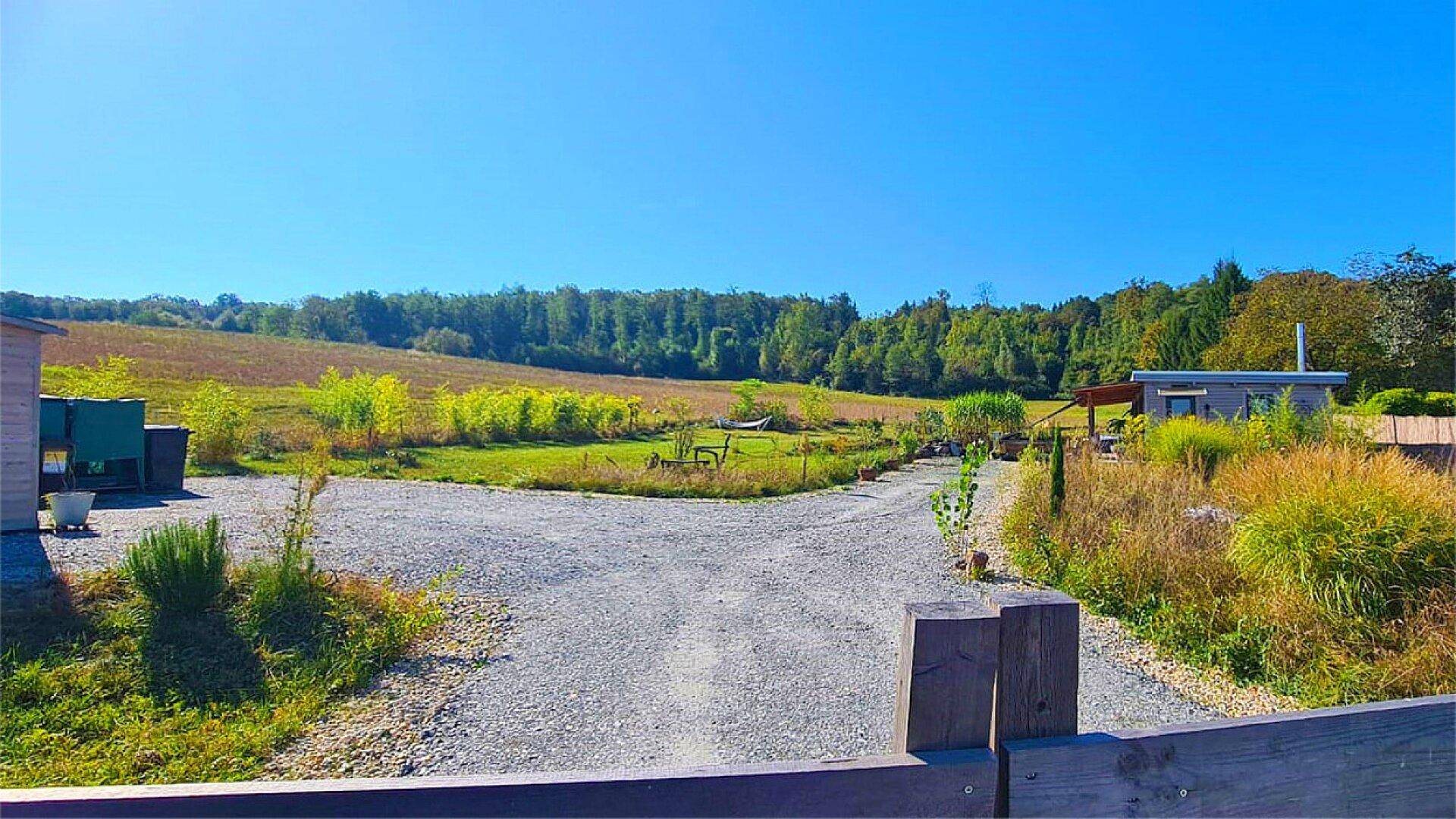 Kiesauffahrt führt durch eine ländliche Umgebung mit Feldern und Wald unter klarem blauem Himmel.