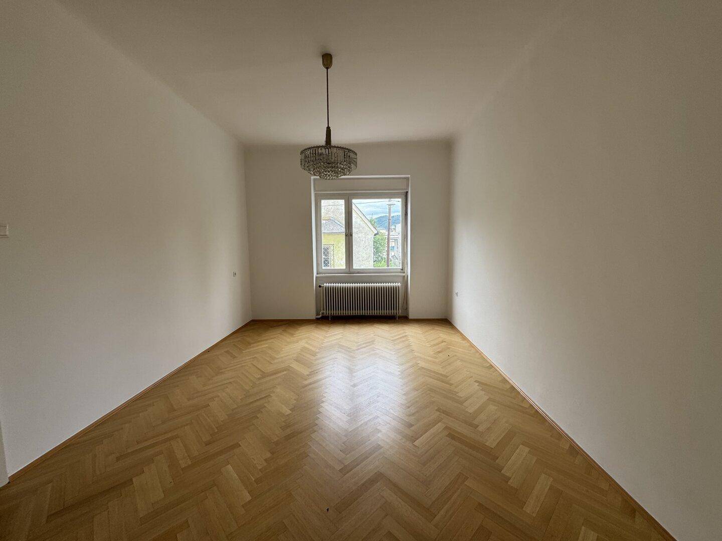Geräumiges Zimmer mit Fischgrätparkett, großem Fenster und Kronleuchter.