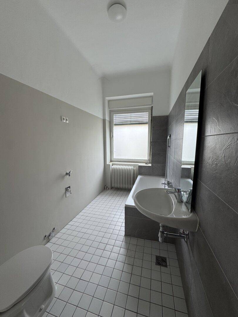 Badezimmer mit grauen Fliesen, Badewanne, Waschbecken und Fenster.