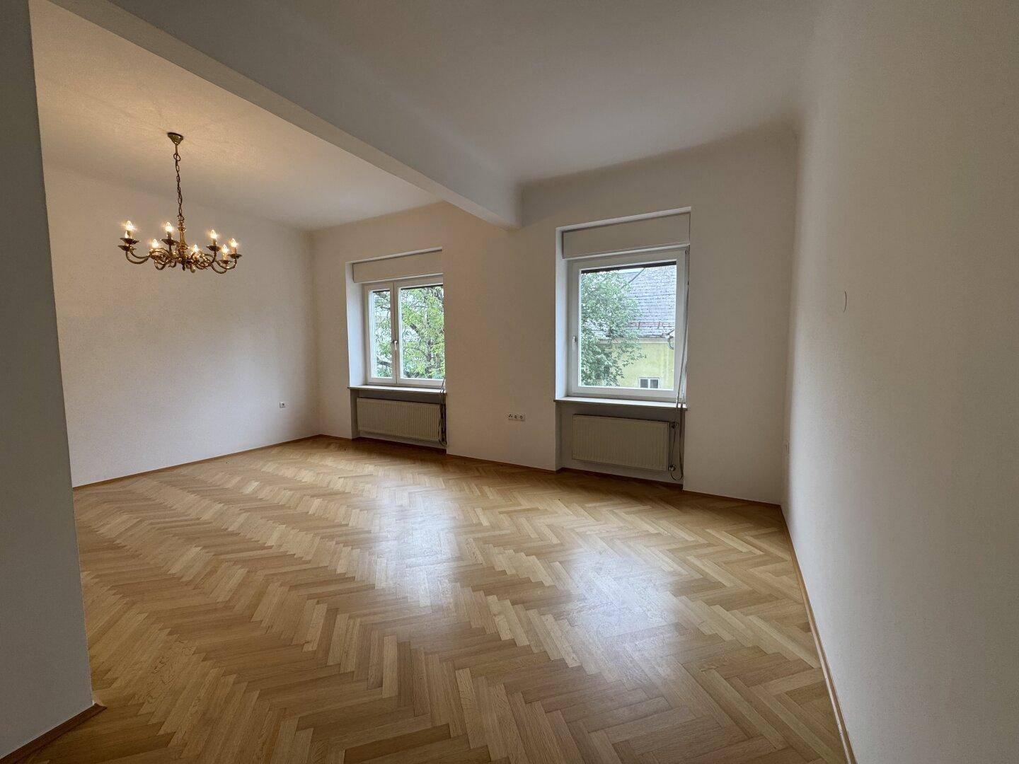 Heller Raum mit Fischgrätparkett, zwei Fenstern und klassischem Kronleuchter.