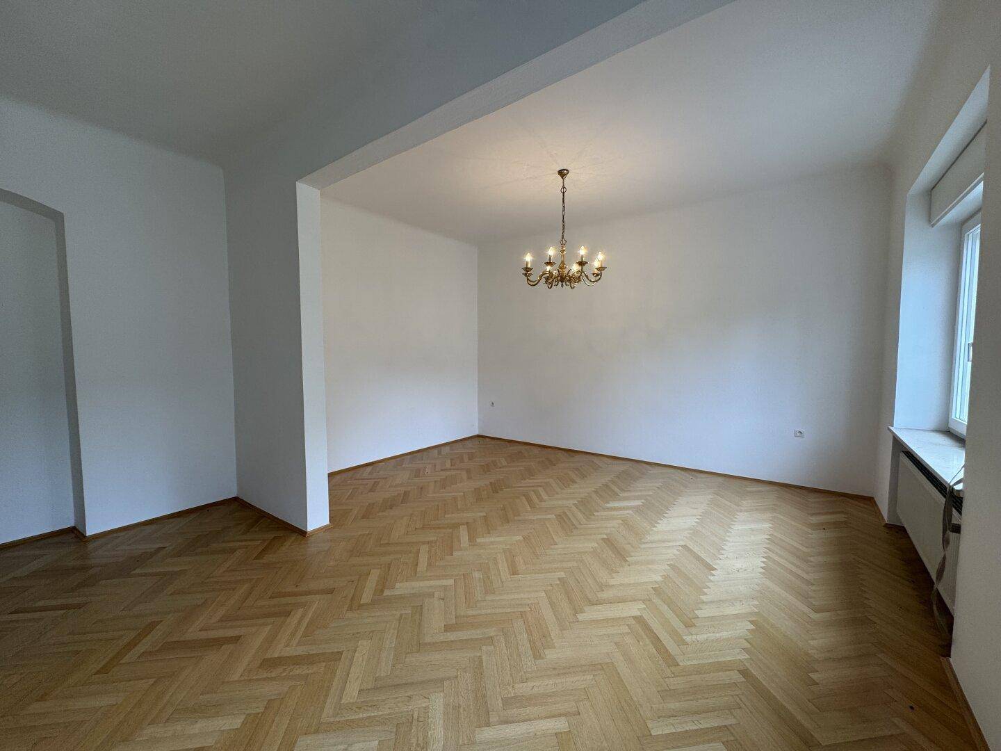 Geräumiger Raum mit Fischgrätparkett, weiß gestrichenen Wänden und Kronleuchter.