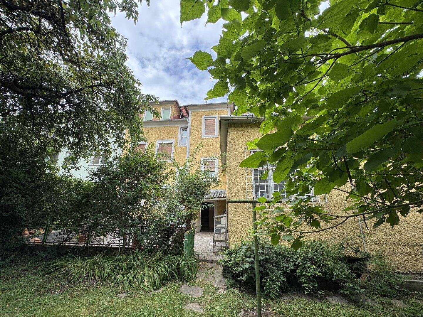 Blick durch Bäume auf die Rückseite des Gebäudes und den Gartenbereich.