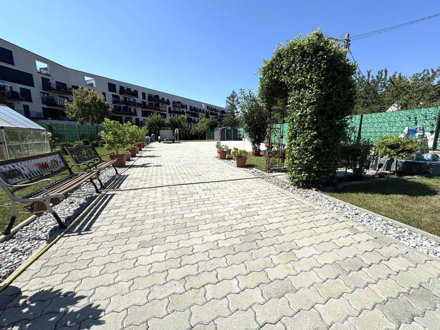 Breiter gepflasterter Weg im Garten mit Sitzbänken und einem begrünten Torbogen.