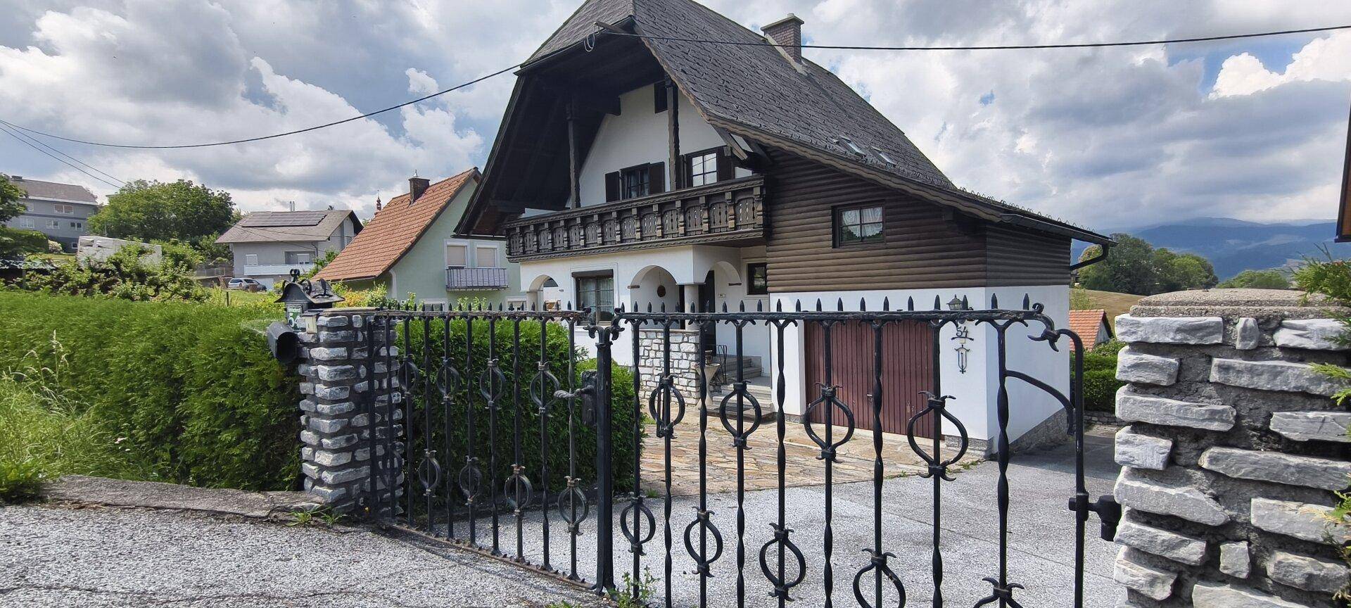 Traditionelles Haus mit schmiedeeisernem Tor und Steinsockel, umgeben von Hecken und Bäumen.