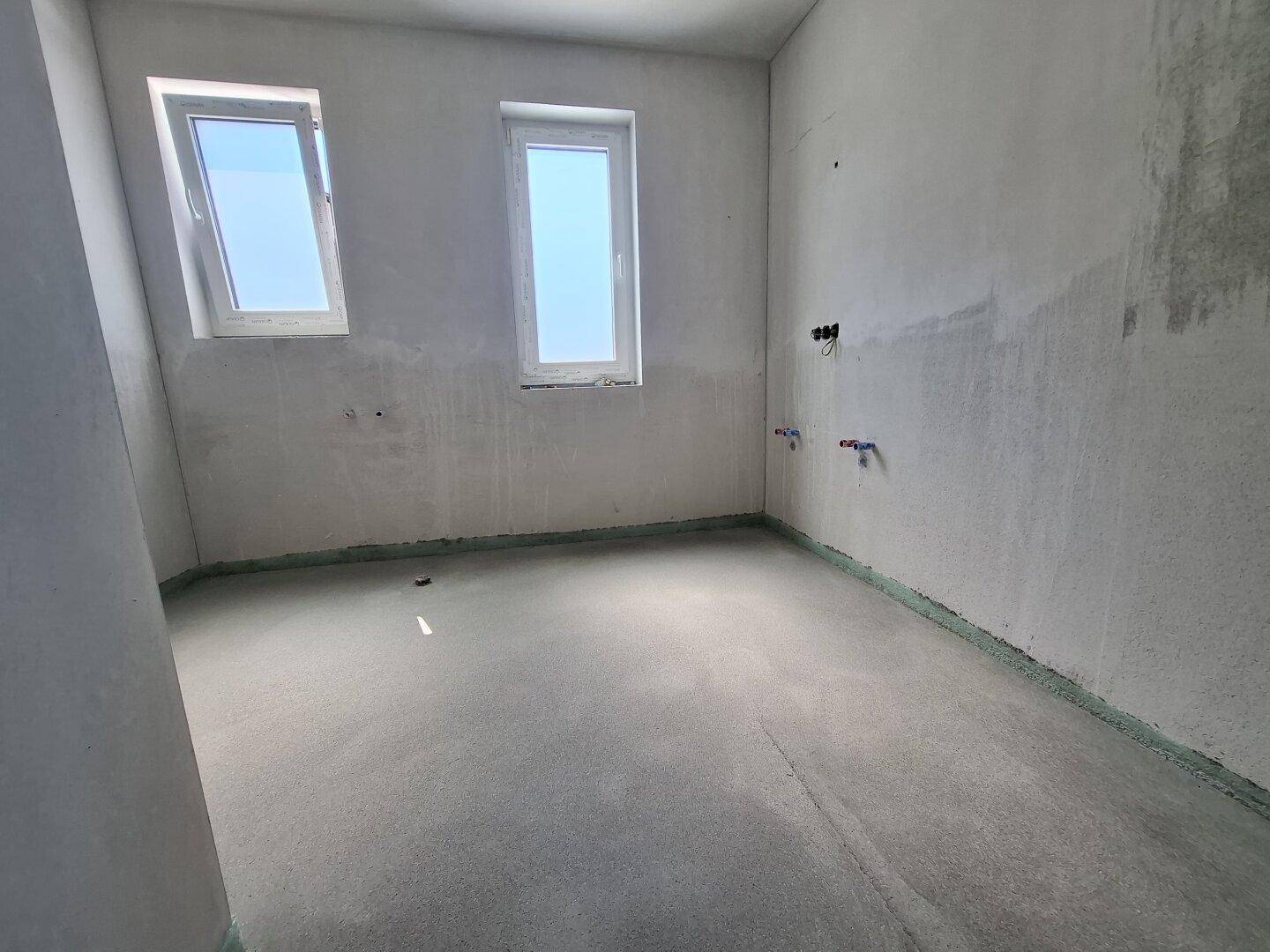 Unfertiges Badezimmer mit zwei Fenstern und vorbereiteten Anschlüssen für Sanitäranlagen.