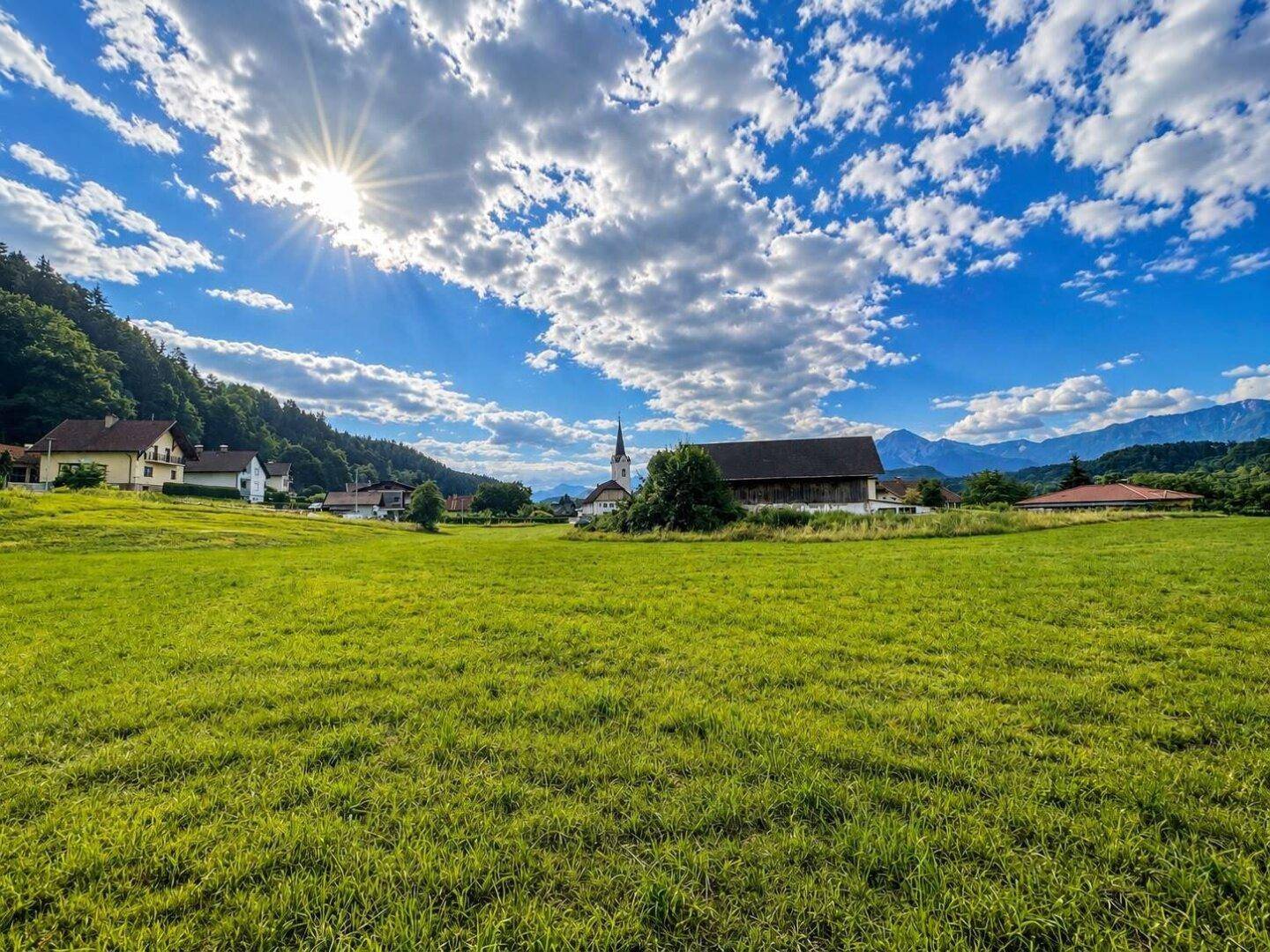 Weitläufige grüne Wiese mit Blick auf ein Dorf, eine Kirche und ferne Berge unter blauem Himmel.