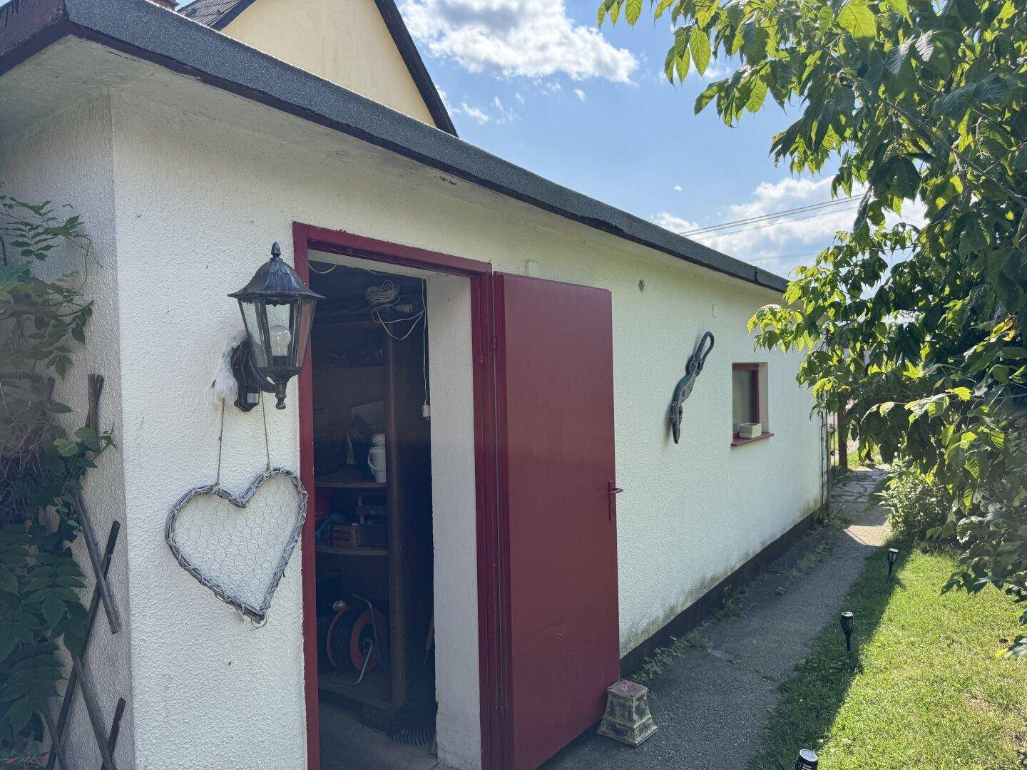 Kleines Nebengebäude mit roter Tür und weißer Fassade im grünen Gartenbereich.