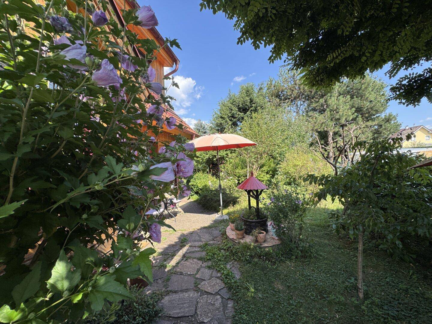 Steinplattenweg führt durch den blühenden Garten mit einem roten Sonnenschirm und Zierbrunnen.