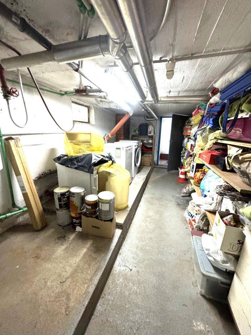 Unaufgeräumter Kellerraum mit Waschmaschine, Trockner und sichtbaren Rohren an der Decke.