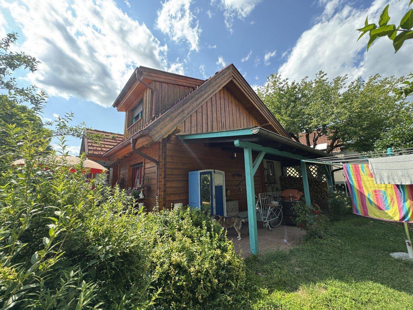 Charmantes Holzhaus mit überdachter Terrasse und üppigem Garten unter blauem Himmel.