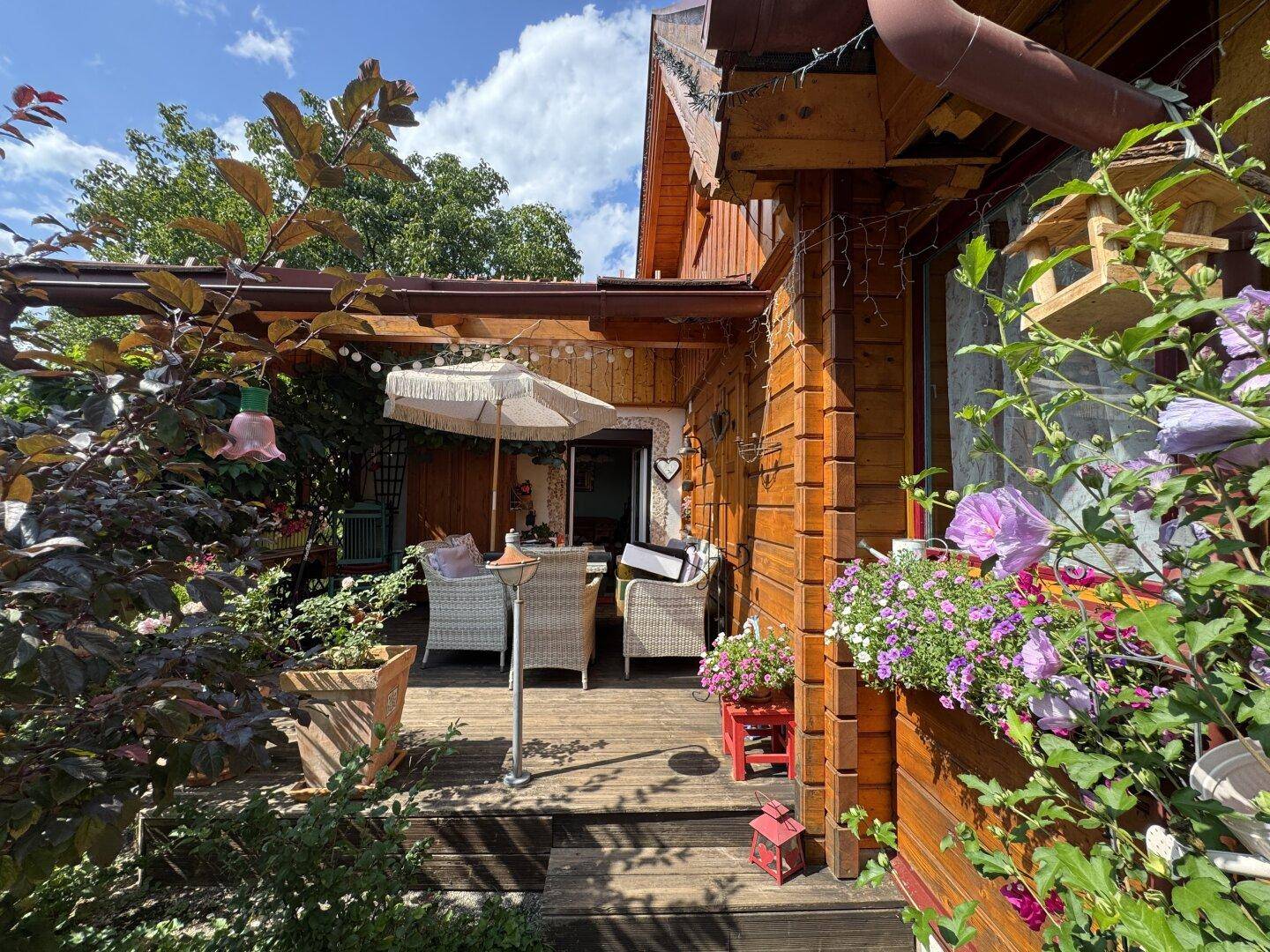 Gemütliche Holzterrasse mit Gartenmöbeln, Sonnenschirm und blühenden Pflanzen in Töpfen.
