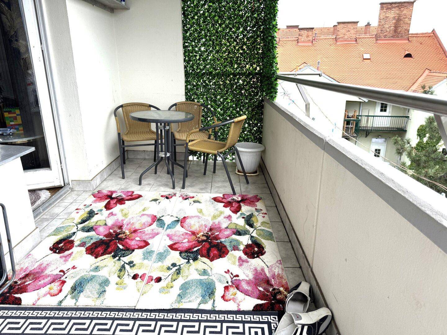 Gemütlicher Balkon mit kleinem Tisch, Stühlen und einer grünen Kunsthecke als Dekoration.