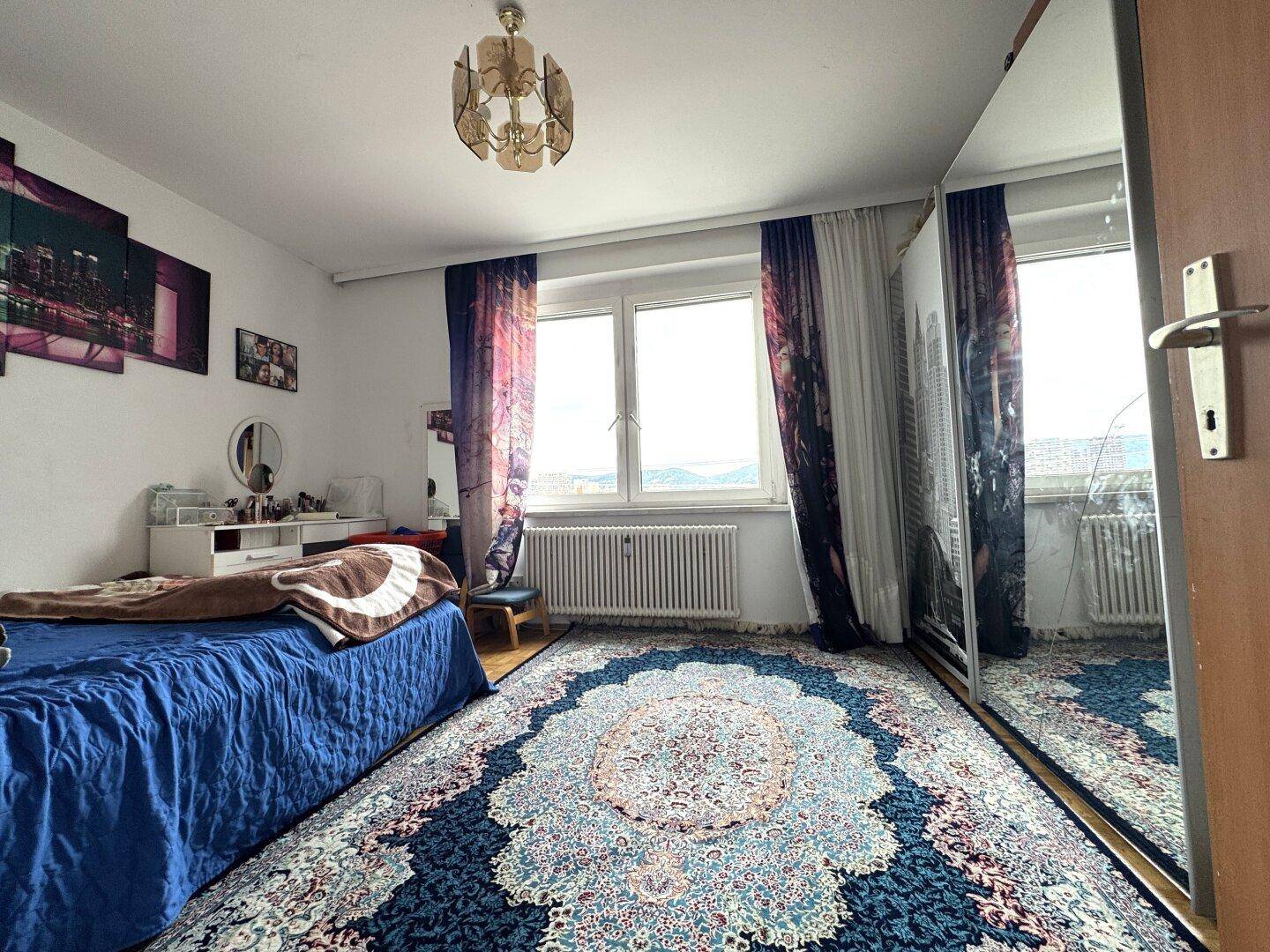 Schlafzimmer mit Bett, großem Spiegelschrank, gemustertem Teppich und Fenster.