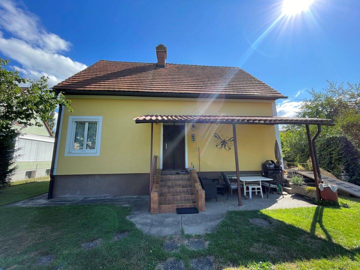 Gelbes Einfamilienhaus mit überdachter Terrasse und kleinem Essbereich im Garten.