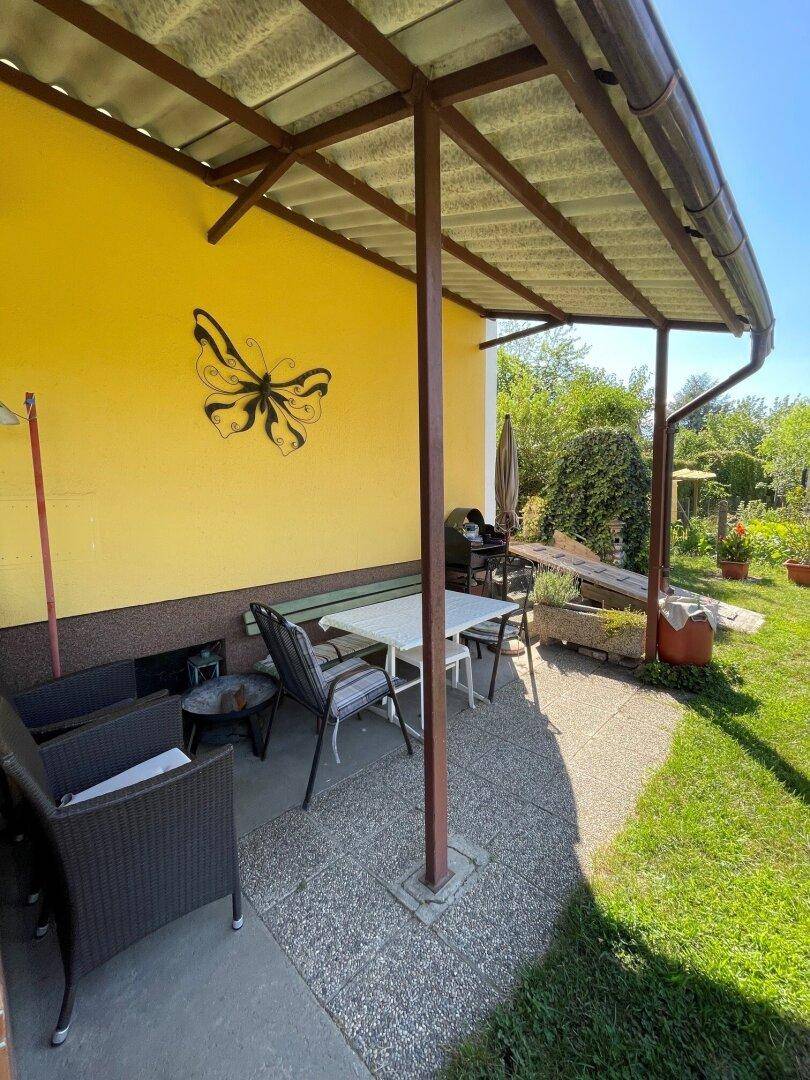 Überdachte Terrasse mit Gartenmöbeln und Schmetterlingsdekoration an der gelben Hauswand.