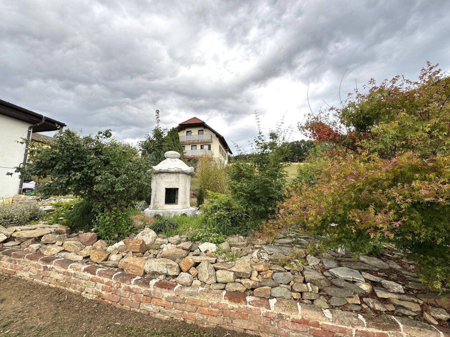 Gartenbereich mit einer Steinmauer, dekorativen Elementen und einem weiteren Haus im Hintergrund.