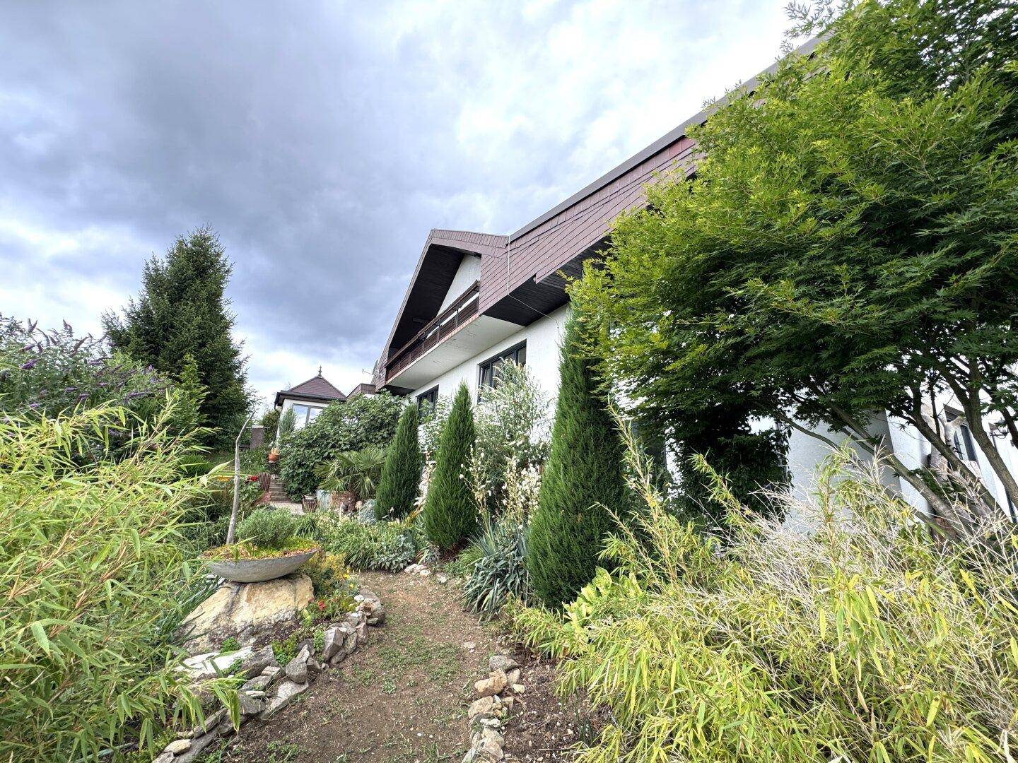 Seitlicher Blick auf das Haus mit einem Gartenweg, verschiedenen Pflanzen und einem Balkon im Obergeschoss.