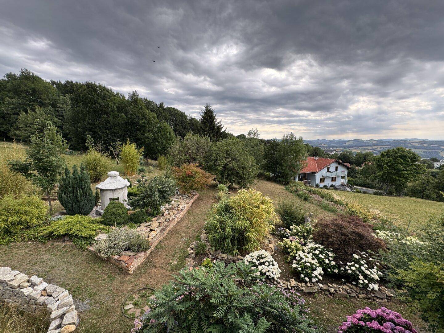 Weitläufiger, gepflegter Garten mit vielfältiger Bepflanzung, Steinmauer und Fernblick auf die Landschaft.