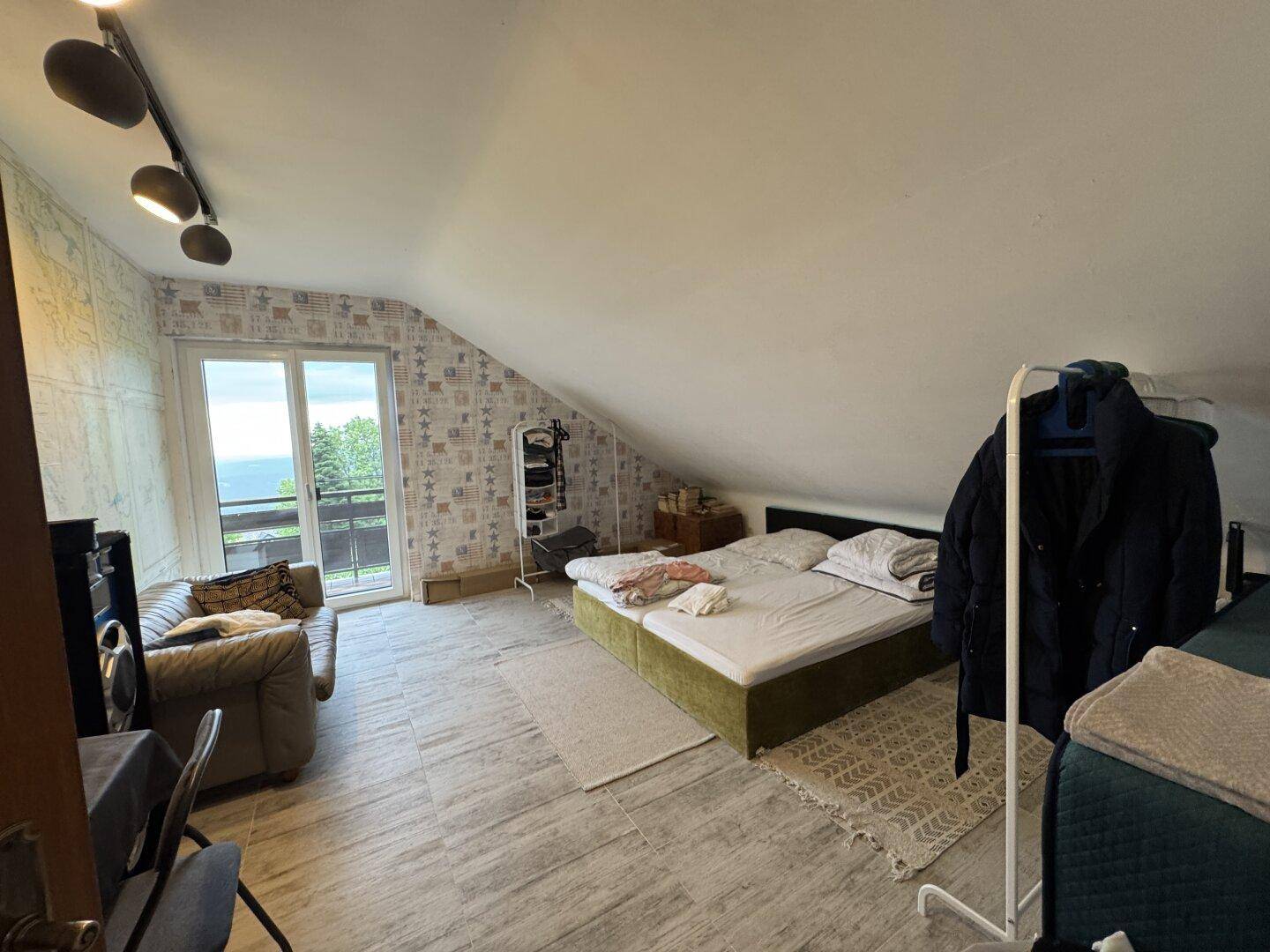 Geräumiges Dachgeschoss-Schlafzimmer mit Fliesenboden, Sofa und Zugang zu einem Balkon mit Weitblick.