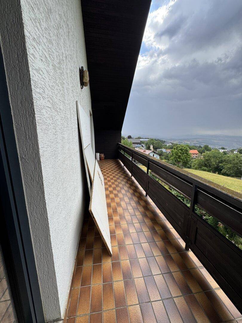 Langer Balkon mit braunen Fliesen und Holzgeländer, bietet einen weiten Blick auf die grüne Landschaft.
