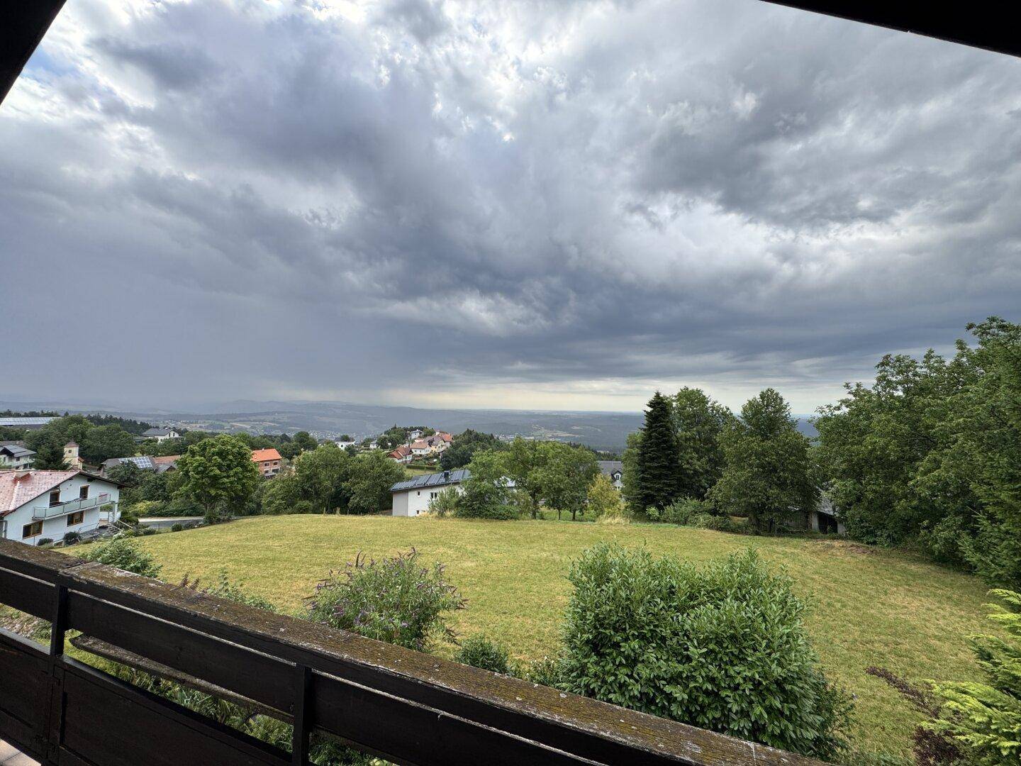 Weitläufiger Blick von einem Balkon über grüne Hügel und eine ländliche Siedlung unter bewölktem Himmel.