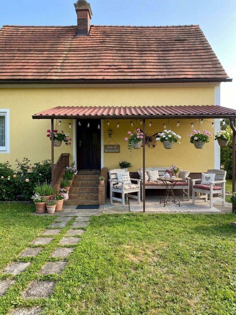 Gemütliche überdachte Terrasse mit Sitzgelegenheiten und Blumenampeln vor dem gelben Haus.